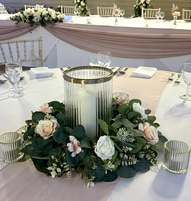 Greenery wreath with pale pink and ivory roses around a glass hurricane vase and candle