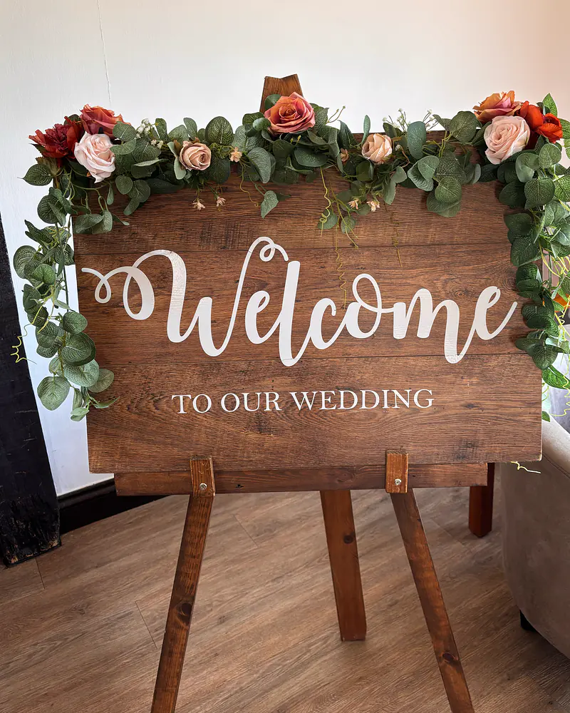 Rustic wooden welcome sign on an easel, dressed with a eucalyptus garland and roses